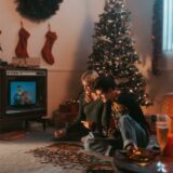 Ein mann und eine frau sitzen in einem weihnachtlich geschmückten wohnzimmer auf dem boden und gucken einen weihnachtsfilm.