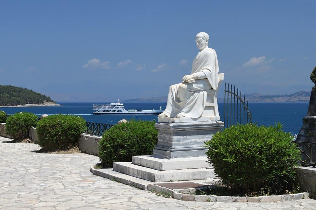 A white marble statue of a seated man in classical attire overlooks a bright blue sea where a large ferry is passing by.