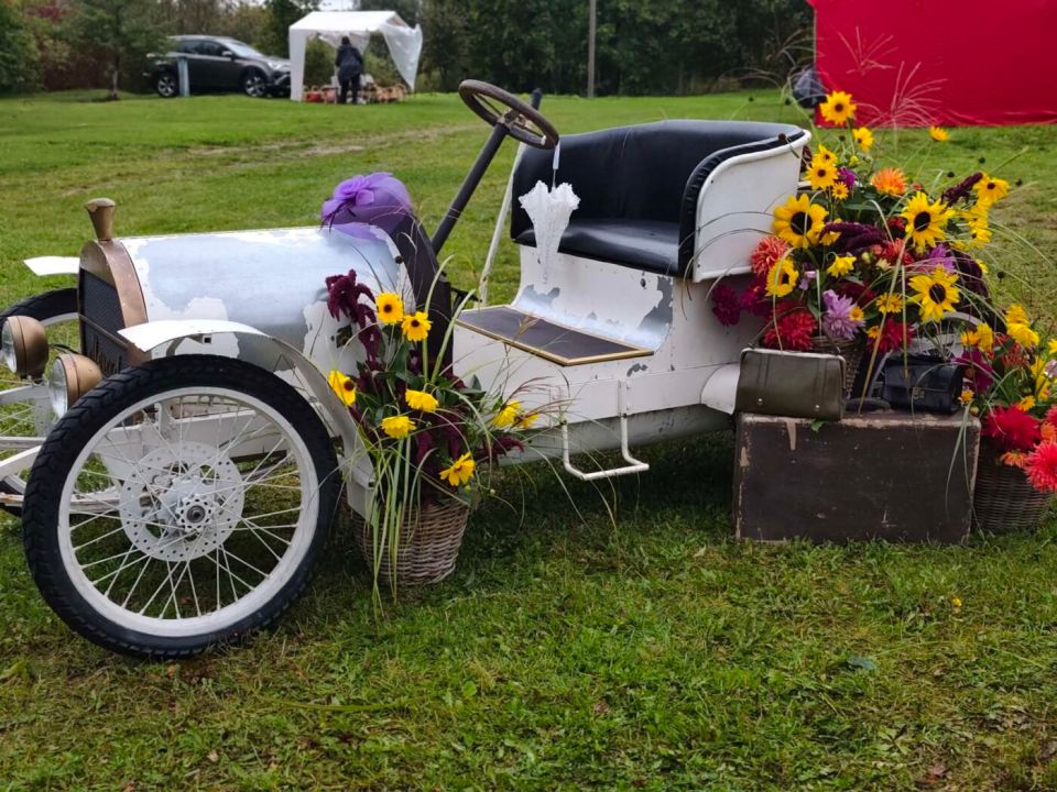 Baltas senovinio automobilio formos eksponatas, papuoštas gėlėmis ir senais lagaminais.