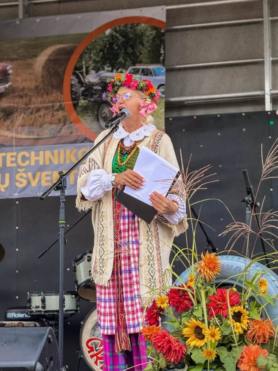 Tautiniais motyvais pasipuošusi šventės vedėja su gėlių vainiku kalba į mikrofoną.