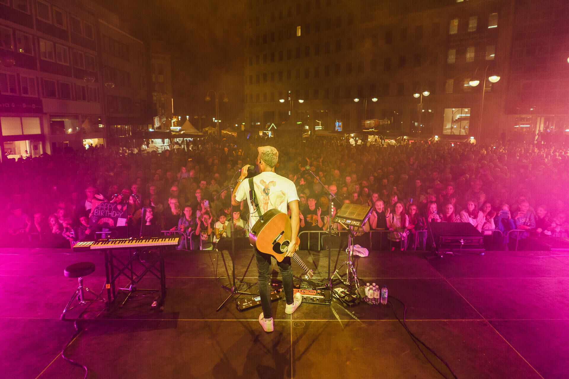 Umsonst und draußen: bochumer musiksommer lockt mit 1000 künstlern 2026 - iustitia. Bg - gerechtigkeit Ein musiker steht mit dem rücken zur kamera, einer gitarre umgehangen und einem mikrofon in der hand auf der bühne vor einem großen publikum.