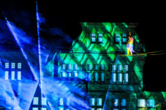 Herbstgenuss: magie aus licht und farbe 2026 - iustitia. Bg - gerechtigkeit Eine artistin balanciert in schwindelerregender höhe auf einem hochseil vor der eindrucksvoll illuminierten fassade eines gebäudes. Sie trägt ein leuchtendes kostüm, das sich farblich von den grün und blau angestrahlten projektionen auf der hauswand abhebt. Lichtstrahlen und nebeleffekte schaffen eine spektakuläre atmosphäre und verleihen der nächtlichen inszenierung eine beinahe magische wirkung. Die kombination aus lichtkunst, architektur und akrobatik macht die szene zu einem eindrucksvollen blickfang.