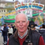 Schausteller frank foulon steht auf einem kirmesplatz und lächelt in die kamera. Im hintergrund sind fahrgeschäfte zu sehen.