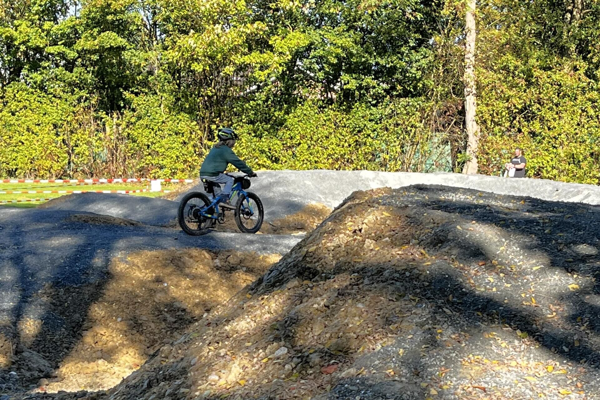 Ein radfahrer fährt mit einem mountainbike über die hügelige strecke einer pumptrackanlage. Die fahrbahn besteht aus dunklem schotter und verläuft in mehreren wellen durch eine grüne umgebung.