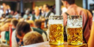 Zwei Bierkrüge stehen auf einer Theke. Im Hintergrund sind viele Menschen in Trachten.