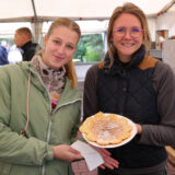 Zwei personen präsentieren auf dem herbstmarkt in nordkirchen gemeinsam einen frisch gebackenen kuchen auf einem teller. Sie stehen lächelnd in einem verkaufszelt, im hintergrund sind weitere menschen und stände zu sehen.